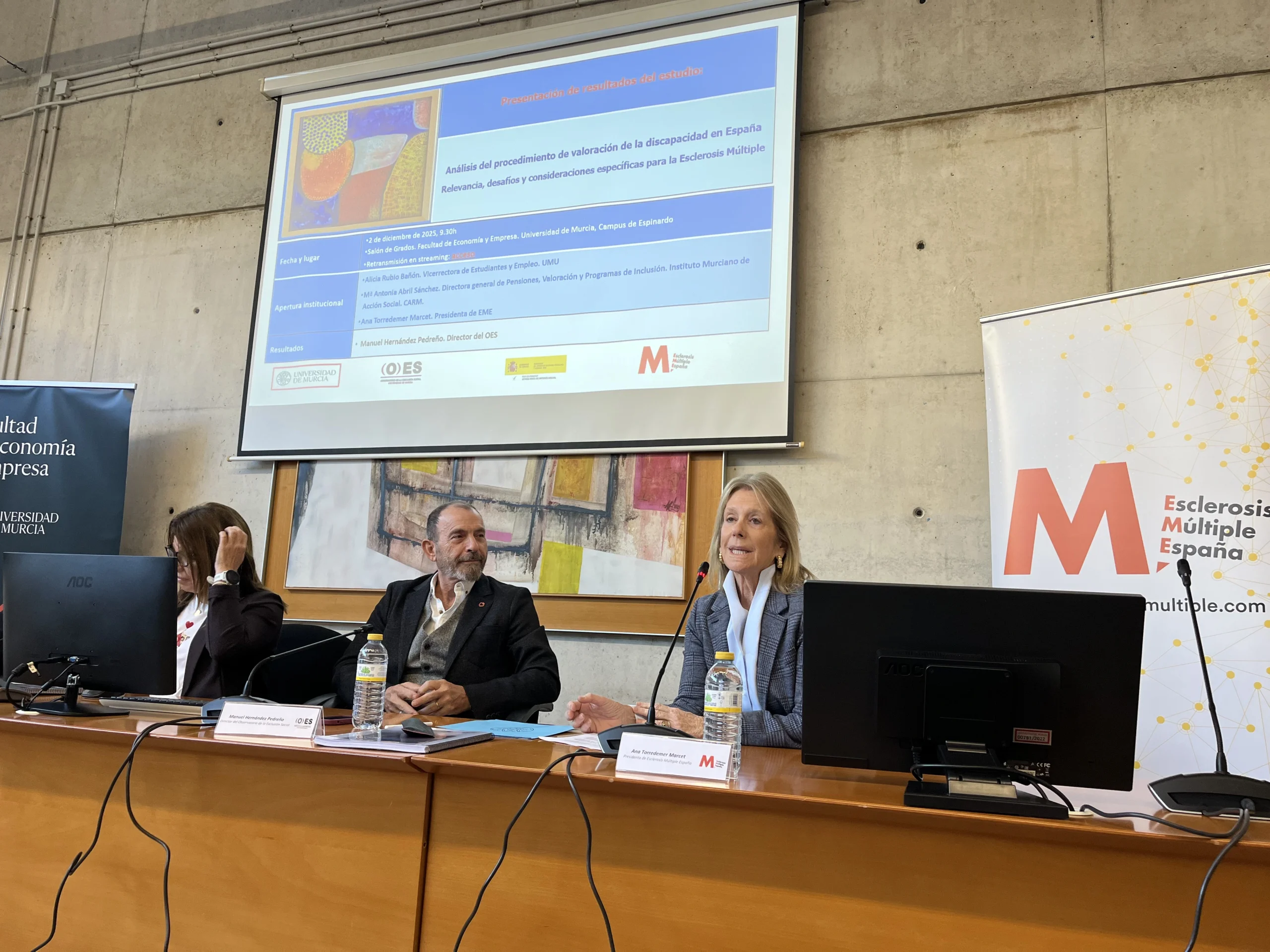 Mesa de presentación del estudio sobre la valoración de la discapacidad en España, con tres ponentes frente al público en la Universidad de Murcia, junto a un panel de Esclerosis Múltiple España.